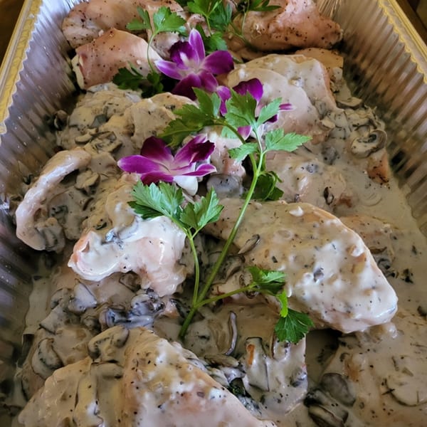 Chicken breasts in creamy mushroom sauce, garnished with parsley and purple orchids in a foil pan.