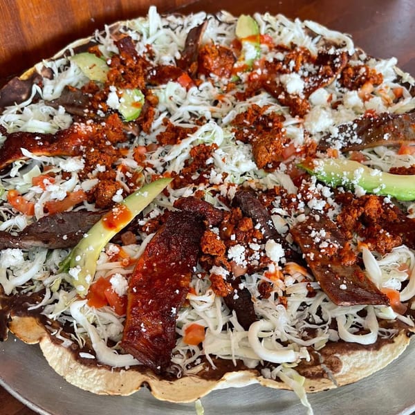 A large, round tlayuda topped with shredded cabbage, chorizo, steak, avocado, and cheese.