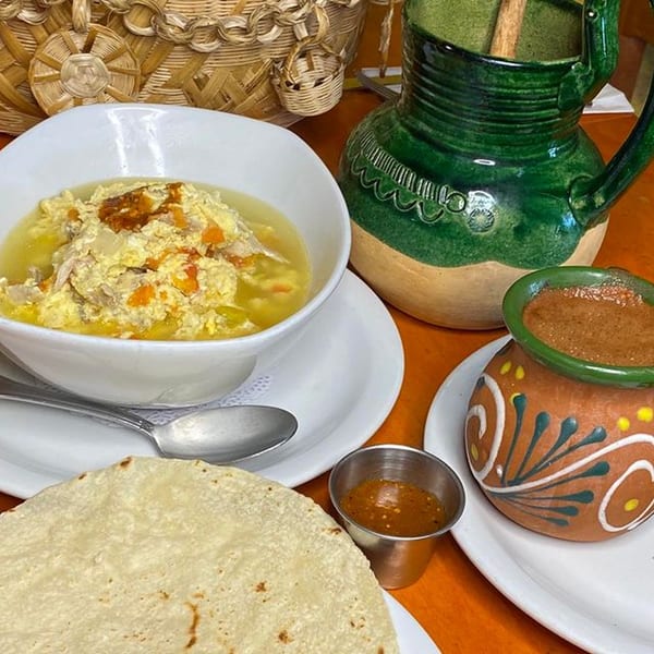 A bowl of Mexican egg soup, tortillas, salsa, and a green ceramic pitcher on an orange table.